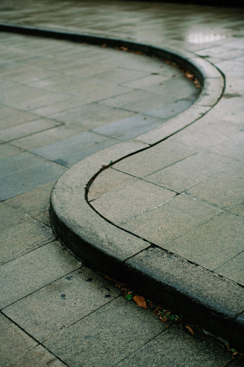 Curved sidewalk with water on wet pavement, evoking subtle leadership, calm orientation, and movement shaped by changing terrain.
