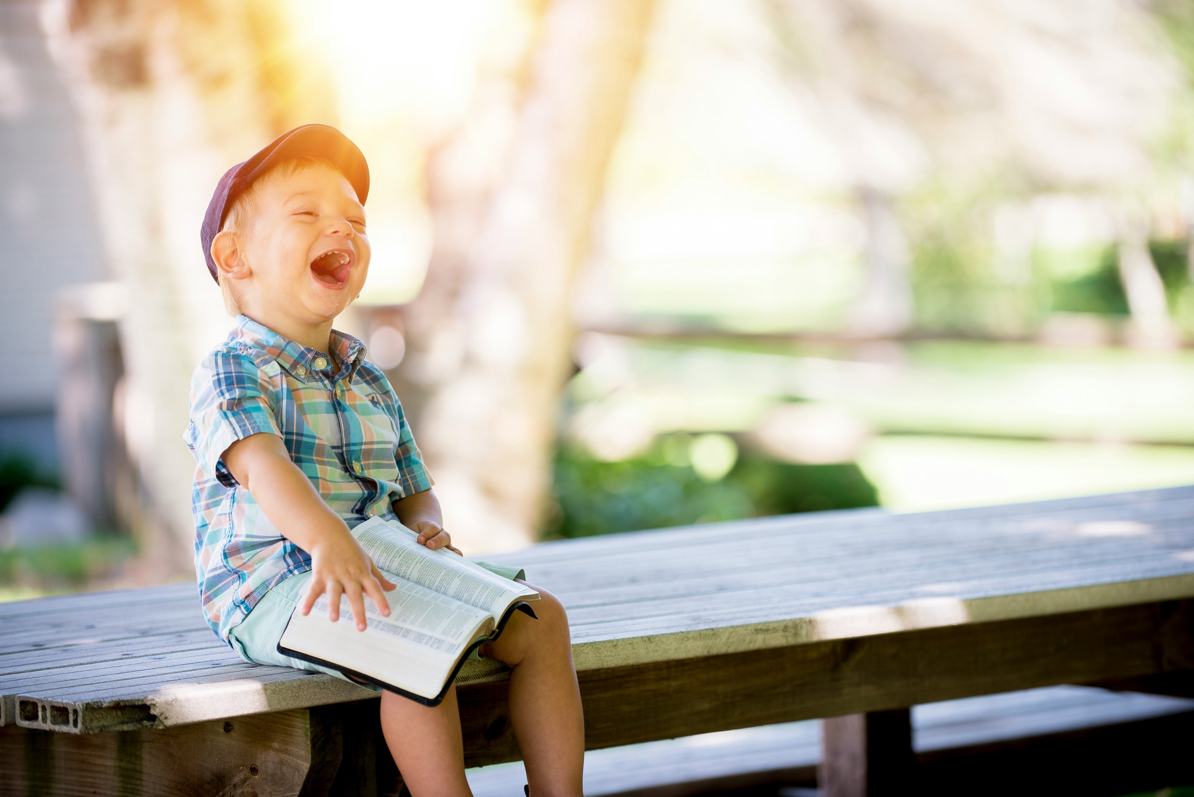 Child sitting on a wooden bench outdoors, holding a book and laughing.