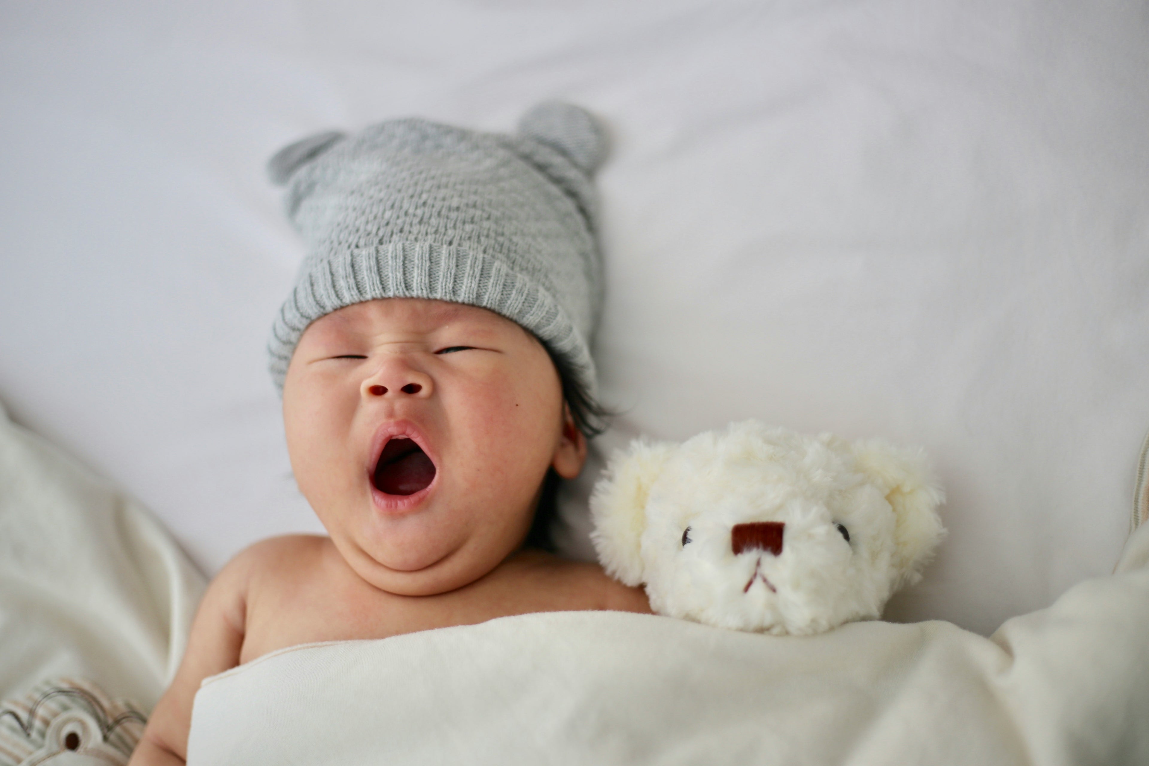 Baby yawning with a gray knit hat and white teddy bear on a white background