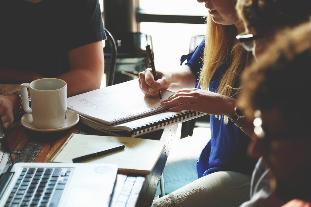 People sitting at a table with notebooks and a laptop, engaged in focused work — a calm symbol of collaboration, shared clarity, and collective intelligence.