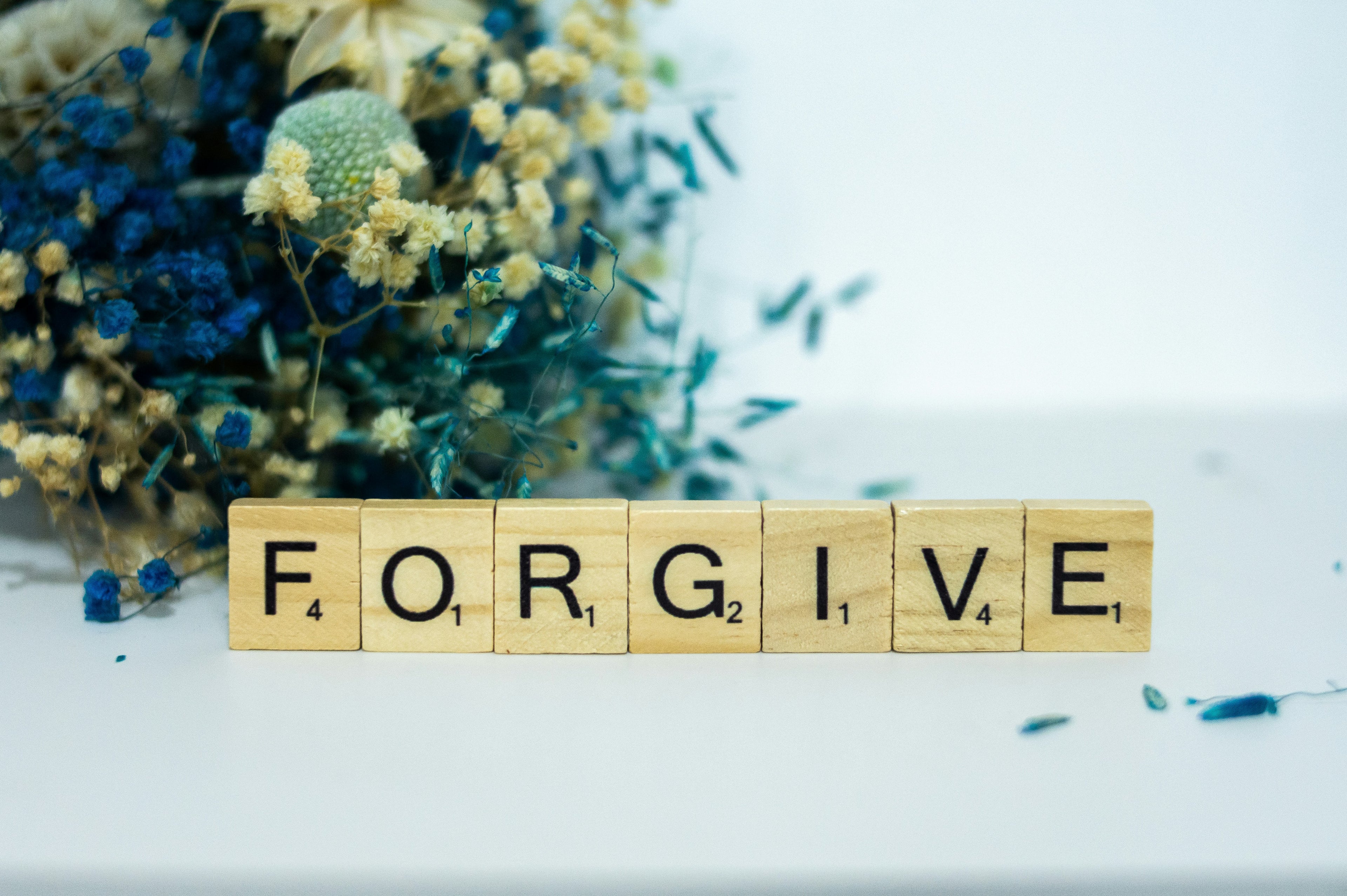 Six wooden Scrabble-style letter blocks spell the word FORGIVE, placed on a white surface with dried flowers in the background.